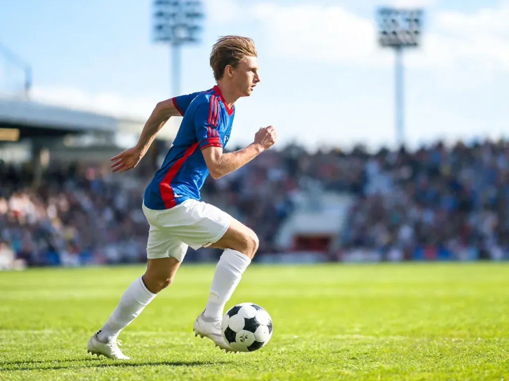 Dembele w niebiesko-czerwonej koszulce biegnie z piłką po boisku, dynamiczna akcja na tle stadionu i kibiców.